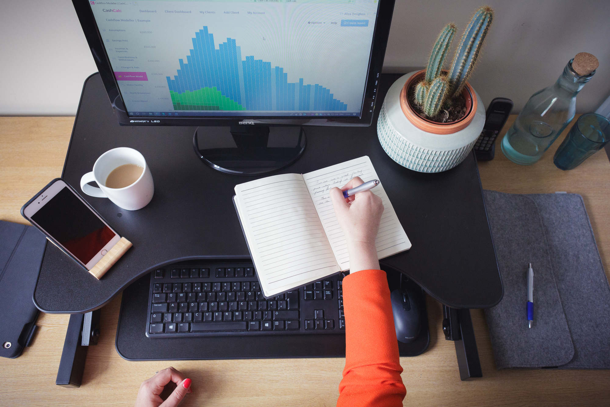 Person sat at desk-writing in notebook-desktop computer-plant-mobile phone-cup of tea