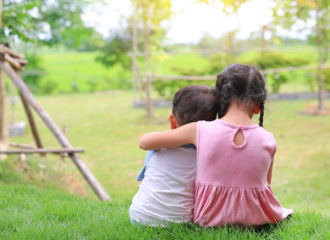 Two small children sitting in the grass-girl's arm is around boy's shoulders