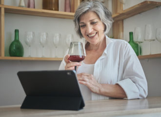 Smiling grey-haired woman using laptop while holding glass of red wine and